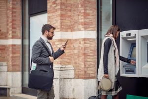 Žena vybírá peníze z bankomatu, muž stojí poblíž