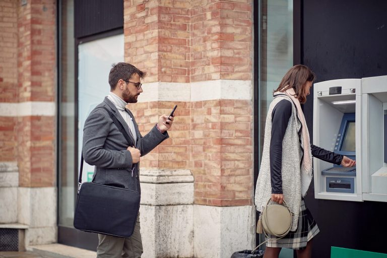 Žena vybírá peníze z bankomatu, muž stojí poblíž